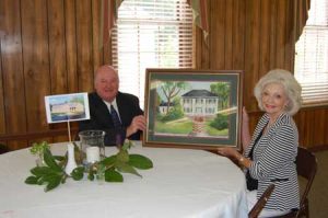 Rev. Barry Cannon and artist Virginia Culbertson hold a picture that Mrs. Culbertson painted of the Shepherd House that sat on the site where the new parsonage is located. The watercolor is painted after an oil painting by Florence Mims Bryan c. 1980. A photo taken by Cynthia Bull of the new parsonage sits on the table beside Rev. Cannon.